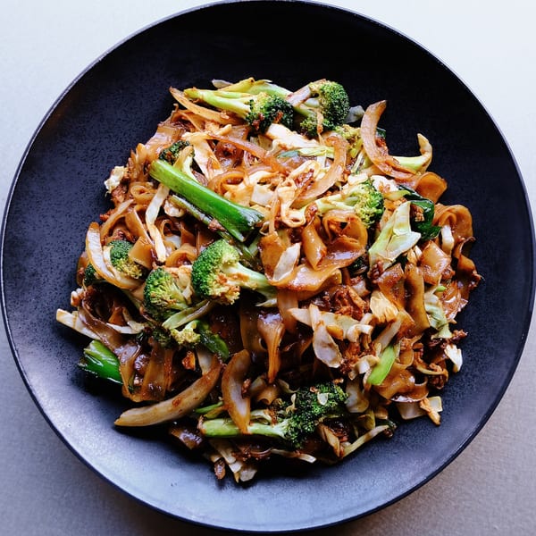 Close-up of Chinese stir-fried noodles with broccoli, cabbage, and onions in a black bowl.