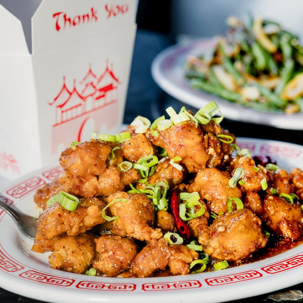 A plate of Chinese orange chicken with green onions, chili peppers, and a takeout box.