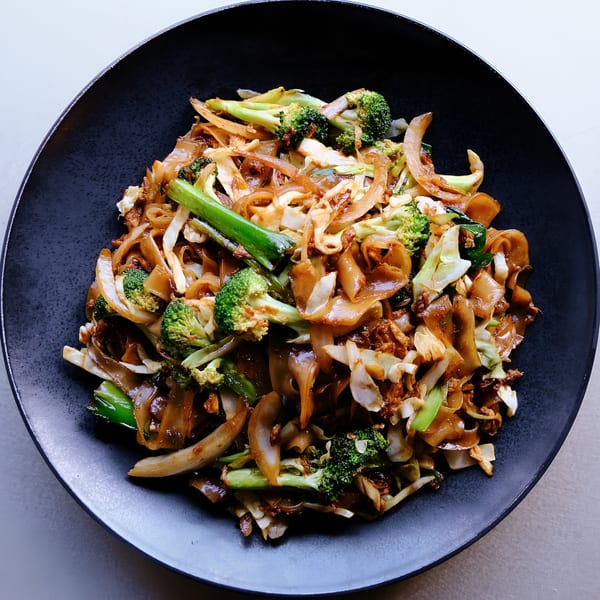 Close-up of Chinese stir-fried noodles with broccoli, cabbage, and onions in a black bowl.