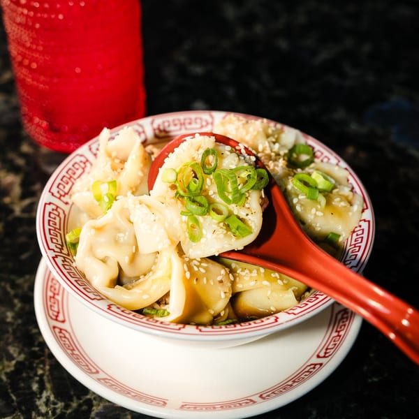 A bowl of Chinese dumplings, garnished with green onions and sesame seeds, with a red spoon.