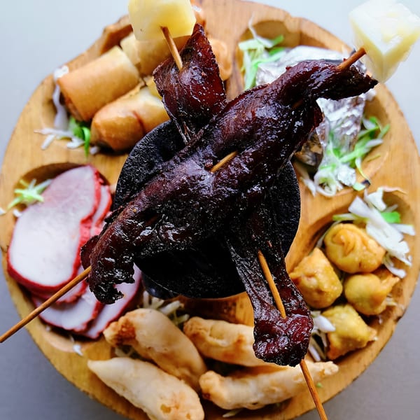 A wooden platter of Chinese appetizers, including spring rolls, char siu, dumplings, and beef skewers.