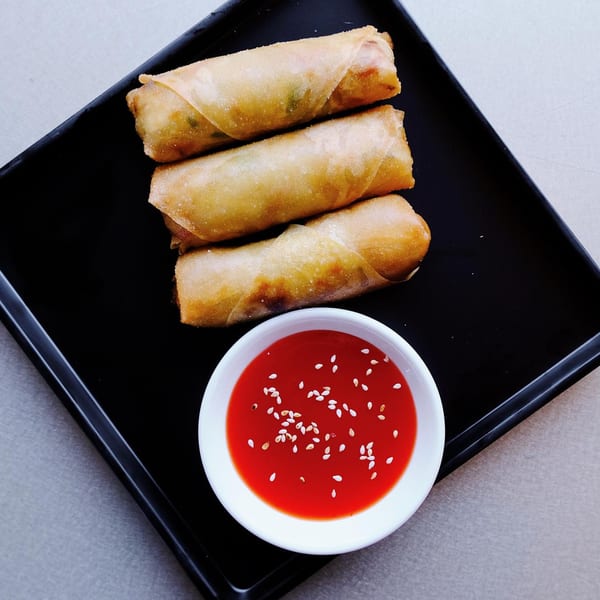 Three golden-brown spring rolls and a small white bowl of red dipping sauce with sesame seeds.