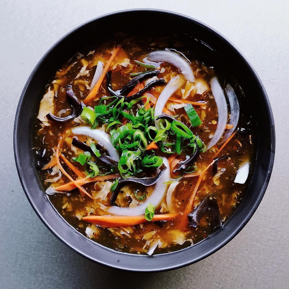 A black bowl of hot and sour soup with carrots, onions, mushrooms, and scallions.